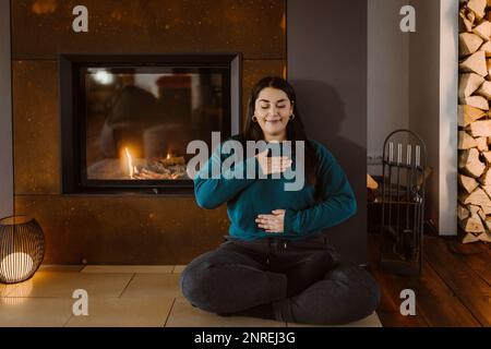 Eine Frau mit geschlossenen Augen, die Atemübungen macht, während sie zu Hause mit gekreuzten Beinen neben dem Kamin sitzt Stockfoto