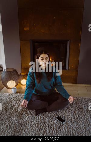 Frau, die Yoga praktiziert, während sie zu Hause mit gekreuzten Beinen auf Teppich sitzt Stockfoto