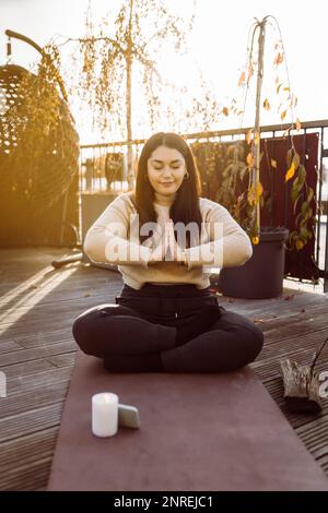 Frau, die Yoga macht, während sie mit gefalteten Händen auf der Trainingsmatte auf der Terrasse sitzt Stockfoto