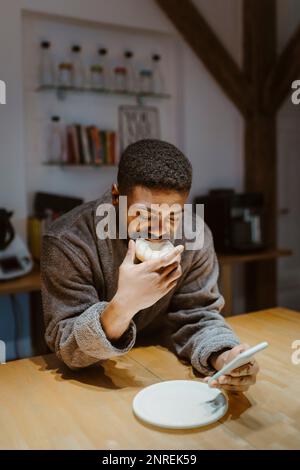 Mann isst Donut und benutzt Smartphone, während er sich zu Hause auf den Tisch lehnt Stockfoto