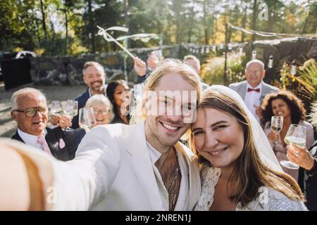 Porträt eines glücklichen frisch verheirateten Paares, das am Hochzeitstag Selfie mit Gästen im Hintergrund macht Stockfoto