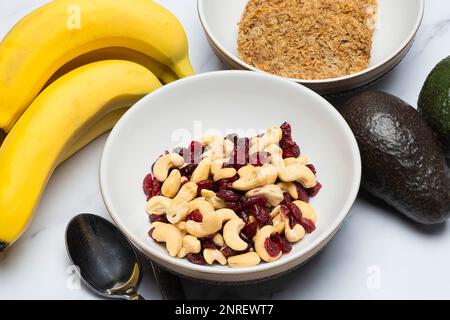 Schale mit Cashews und Cranberrys neben Bananen-Avocados und eine Schüssel mit zwei Frühstückskeksen Stockfoto