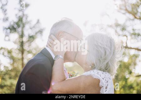 Romantische frisch verheiratete Senior Braut küsst Bräutigam auf dem Mund während der Hochzeit an sonnigen Tagen Stockfoto