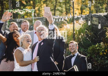 Glücklicher Senior-Mann, der während der Hochzeitszeremonie Selfie mit Braut und Familie auf dem Smartphone macht Stockfoto