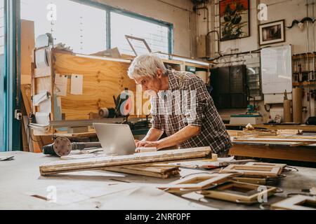 Fokussierter Senior-Zimmermann, der in der Werkstatt an einem Laptop arbeitet Stockfoto