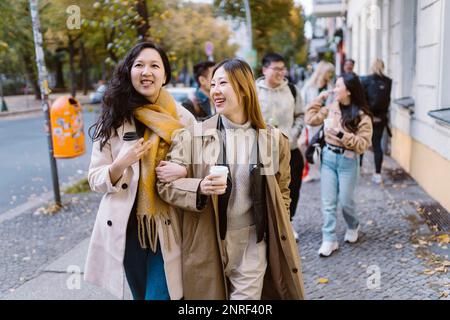 Lächelnde weibliche Freunde, die mit Arm in Arm auf dem Bürgersteig gehen Stockfoto