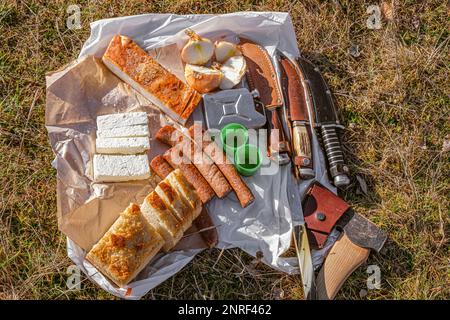 Traditionelles rumänisches Essen mit Würstchen, Zwiebeln und Käse, Schinken und Paprika sowie einer Sammlung von Messern und einer Axt Stockfoto