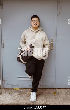 Lächelnder junger Mann mit der Hand in der Tasche, der sich an die graue Tür lehnt Stockfoto