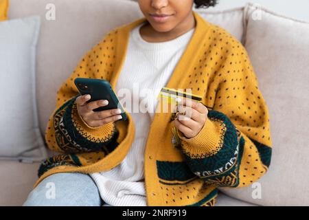 Frau kauft online mit Smartphone und bezahlt mit goldener Kreditkarte. afroamerikanisches Mädchen, das im Internet kauft, Kreditkartendetails eingeben. E-Commerce-Lieferservice für Online-Shopping Stockfoto