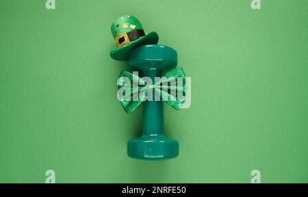 Gym-Kurzhantel mit irischem Shamrock-Kleeblatthut und grüner Fliege. Gesundes Fitness-Flat-Lay-Konzept für St. Patricks Tag. Stockfoto
