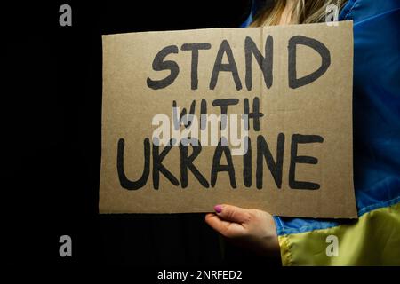 Frau mit ukrainischer Flagge und Hinweisschild auf dem ukrainischen Plakat als Protestmanifestation gegen die russische Invasion und den Krieg gegen die Ukraine. Stockfoto