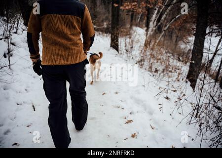 Ein Mann in einem orangefarbenen Sweatshirt, der seinen braunen Hund auf einem schneebedeckten Pfad führt Stockfoto
