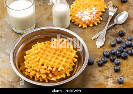 Wiener Waffeln in Metallschüssel. Waffeln und Beeren, Löffel und Gabel auf dem Tisch. Milch in Glas und Flasche. Draufsicht. Rostiger Hintergrund. Stockfoto