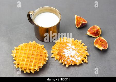 Wiener Waffeln und Feigenhälften auf dem Tisch. Milch in einem Metallbecher. Draufsicht. Grauer Hintergrund. Stockfoto