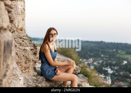 Eine Frau in Jeansanzug sitzt in der Nähe der Steinmauer auf einem Berg in der Nähe der Stadt Stockfoto