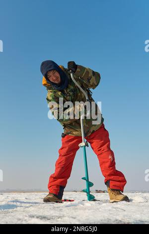 Ein warm gekleideter Asiate bohrt im Winter ein Loch ins Eis Stockfoto