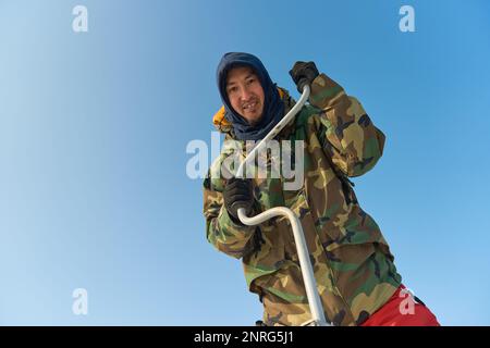 Ein warm gekleideter Asiate bohrt im Winter ein Loch ins Eis Stockfoto