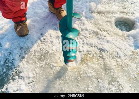 Ein warm gekleideter Asiate bohrt im Winter ein Loch ins Eis Stockfoto