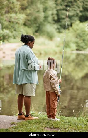 Vertikales Rückansicht-Porträt von Mutter und Tochter, die gemeinsam am Fluss fischen und die Natur genießen Stockfoto