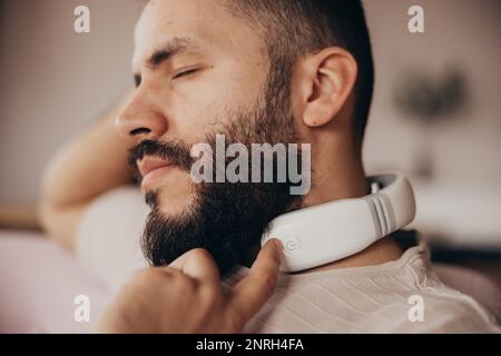 Der junge Mann benutzt ein Massagegerät für den Nacken, um sich zu Hause zu entspannen Stockfoto