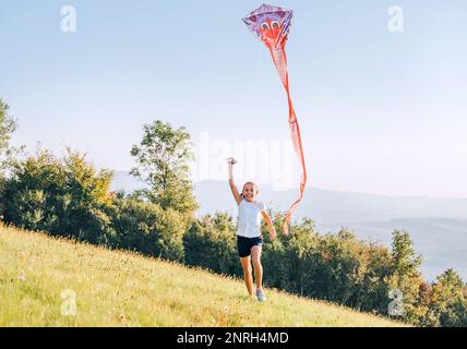 Lächelndes Mädchen mit einem fliegenden bunten Drachen, der auf einer grünen Wiese in den Bergfeldern rennt. Glückliche Kindheitserlebnisse oder Zeit im Freien Stockfoto