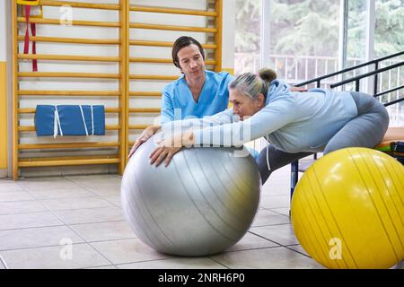 Seniorin mit Physiotherapeut-Anleitung beim Training auf Fitnessball im Reha-Zentrum Stockfoto