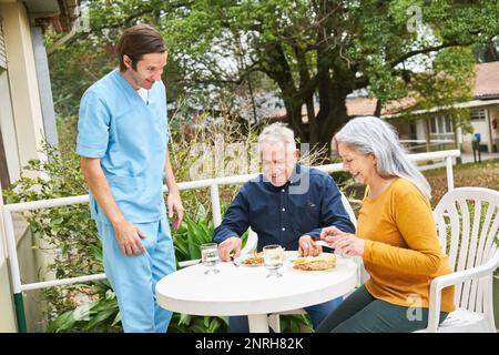 Männliche Krankenschwester schaut auf glücklichen älteren Mann und Frau, die am Tisch im Garten des Pflegeheims essen Stockfoto