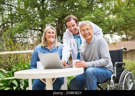 Portrait eines glücklichen Pflegers, der mit einem älteren Paar über einen Laptop spricht, während er im Garten des Pflegeheims Kaffee trinkt Stockfoto