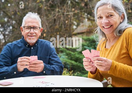 Porträt von Seniorenmann und Seniorenfrau beim Kartenspielen am Tisch im Garten des Pflegeheims Stockfoto