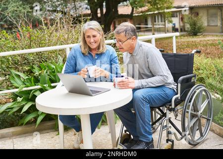 Glücklicher Seniorenmann im Rollstuhl, der mit einer Frau über Laptop am Tisch im Garten des Pflegeheims spricht Stockfoto