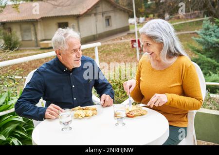 Glücklicher Seniorenmann und glückliche Seniorenmahlzeit am Tisch im Garten des Pflegeheims Stockfoto