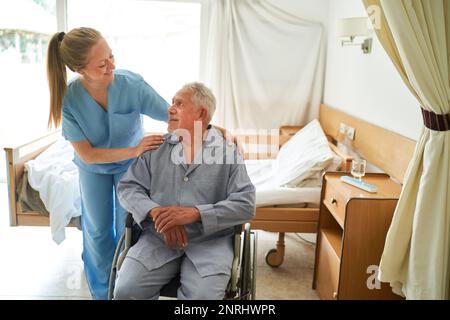 Lächelnde Pflegekraft mit Händen an der Schulter eines im Rollstuhl sitzenden Seniorenmannes im Reha-Zentrum Stockfoto
