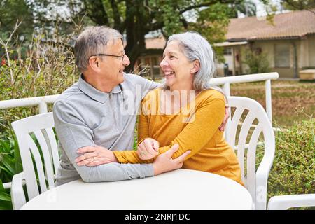 Glückliche Seniorin und umarmter Mann, während sie im Garten des Pflegeheims am Tisch sitzen Stockfoto