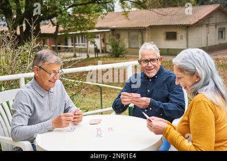 Glückliche Seniorenfreunde, die Karten auf dem Tisch im Garten des Pflegeheims spielen Stockfoto