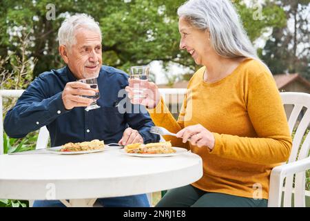 Glücklicher Seniorenmann und glückliche Frau, die ein Glas Wasser toasten, während sie am Tisch im Garten des Pflegeheims essen Stockfoto