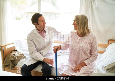 Lächelnder männlicher Arzt, der mit einer älteren Frau im Bett im Reha-Zentrum sitzt Stockfoto