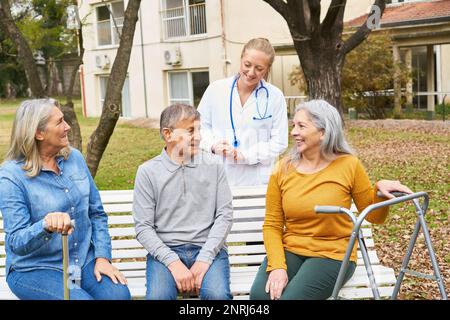 Ein Arzt, der mit einer glücklichen Seniorengruppe spricht, die auf einer Bank im Garten des Pflegeheims sitzt Stockfoto