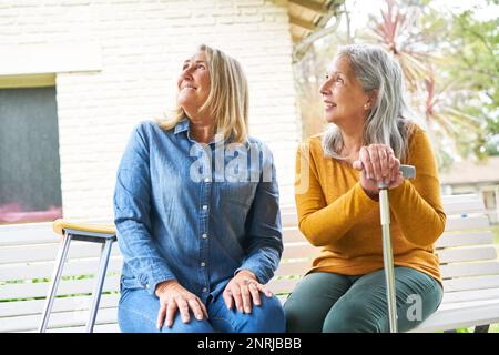 Lächelnde ältere Frauen, die aufblickten, während sie mit Knirschen und Gehstock auf einer Bank im Garten saßen Stockfoto