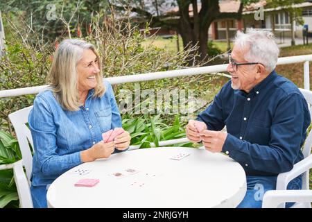 Glücklicher alter Mann und glückliche Frau, die Karten spielt, während sie im Garten des Pflegeheims am Tisch sitzt Stockfoto