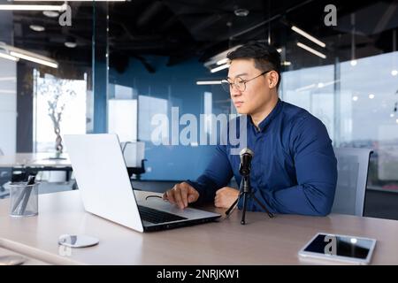 Seriöser Lehrer, der einen Online-Business-Kurs am Arbeitsplatz im Büro aufnimmt, asiatischer Mann mit Laptop, der Schüler online unterrichtet, Geschäftsmann in lässigem Hemd. Stockfoto