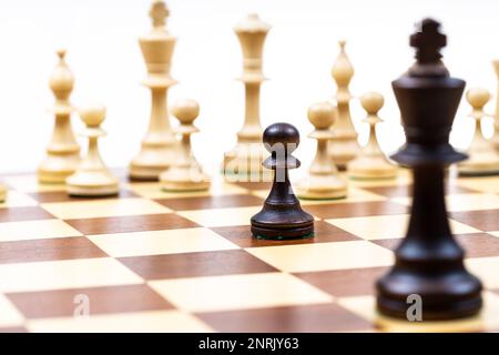 Schwarzer Bauer und einzelne Königin gegen weiße Schachfiguren im Hintergrund auf Holz-Schachbrett Nahaufnahme (Fokus auf den schwarzen Bauern) Stockfoto