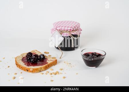 Traditioneller Marmeladentopf mit saurem Kirschrot und leerem Etikett, einer kleinen Schüssel und einer Scheibe Brot mit isolierter Marmelade auf weißem Hintergrund. Stockfoto