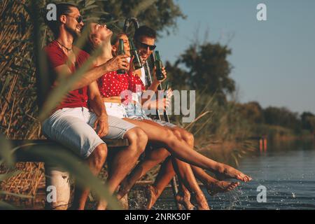 Gruppe junger Menschen, die auf einer Holzplattform am Fluss sitzen, Bier trinken und an warmen Sommertagen ihre Füße ins Wasser spritzen