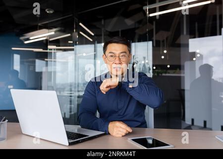 Erfolgreicher Geschäftsmann im Büro, der die App auf einer Smartwatch verwendet, um zu kommunizieren, asiatischer Mann, der künstliche Intelligenz über eine Uhr Sprachbefehle gibt, Chef, der mit einem Laptop arbeitet. Stockfoto