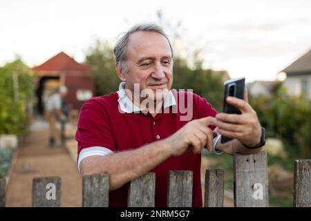 Ein erwachsener Mann, der entspannt neben dem Zaun steht und im Garten telefoniert und lächelt Stockfoto
