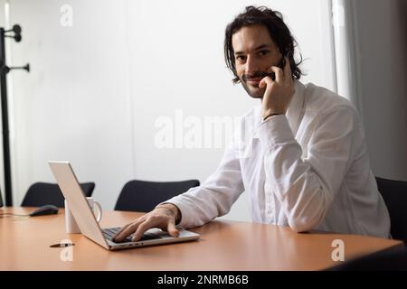 Porträt eines jungen Geschäftsmanns oder Büromitarbeiters, der mit seinem Laptop in einem gemütlichen Büro arbeitet und mit seinem Mobiltelefon kommuniziert Stockfoto