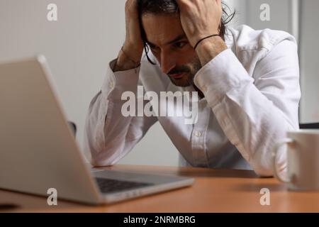 Porträt eines jungen Büroangestellten, der vor seinem Computer sitzt. Er ist müde und gestresst und hält seinen Kopf in den Händen Stockfoto