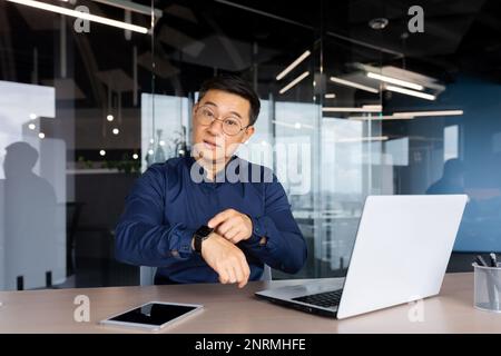 Mann bei der Arbeit mit Laptop im Büro, Geschäftsmann unglücklich und ernst, zeigt Uhr vor der Kamera und verspätet sich zum Meeting eines Kollegen. Stockfoto