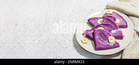 Dünne lavendelfarbene Pfannkuchen mit Bananenscheiben und süßem Belag. Gesundes veganes Lactose- und glutenfreies Essen, Frühstückskonzept. Trendiges, hartes Licht, Dar Stockfoto
