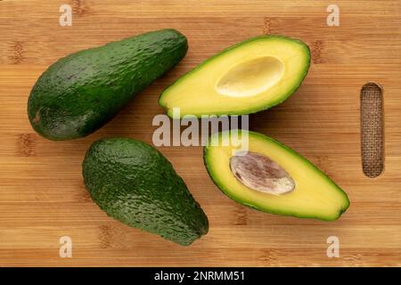 Zwei ganze und zwei halbierte organische Avocados auf einem Bambusschneidbrett, Makro, Draufsicht. Stockfoto